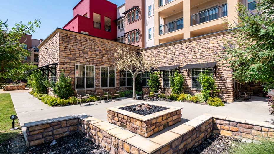 A courtyard with a stone fire pit in the center, surrounded by stone walls and benches. There are several potted plants and small trees. The background features a multi-story building with red, beige, and brown facade and balconies.