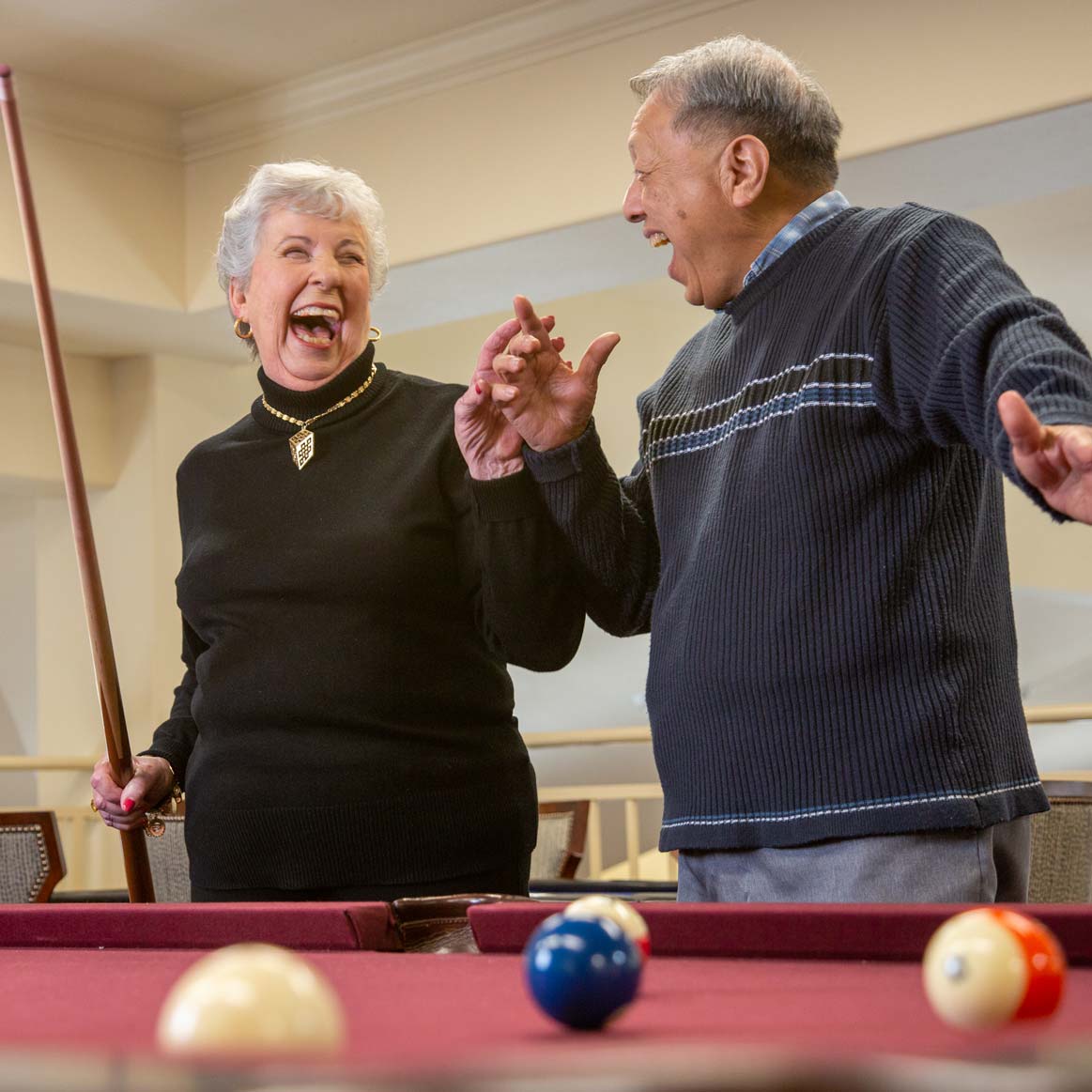 An elderly woman and man share a laugh while standing near a pool table, holding pool cues. Colorful billiard balls are scattered on the table in the foreground. Both are casually dressed and appear to be enjoying a lighthearted moment.