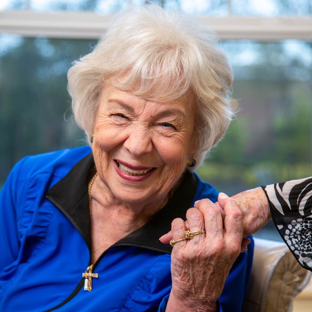 An elderly woman with short white hair, wearing a blue jacket and a gold cross necklace, smiles warmly while holding the hand of another person. They are indoors, with a window showing a blurred outdoor scene in the background.