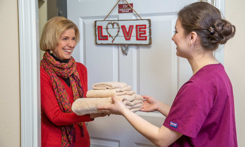 A smiling woman wearing a red sweater and scarf hands a stack of folded towels to another woman dressed in a purple uniform. They stand in front of a door with a decorative sign reading 