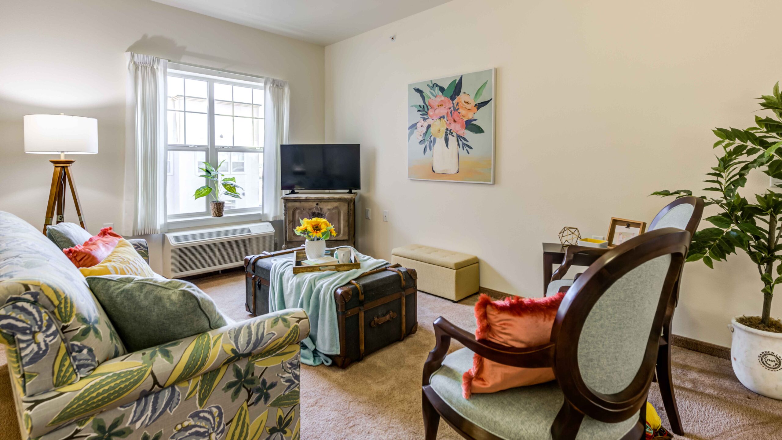 Bright living room with a floral sofa, two chairs, TV, large window with white curtains, potted plants, a trunk-style coffee table, and floral artwork on the wall. The space feels cozy and inviting.