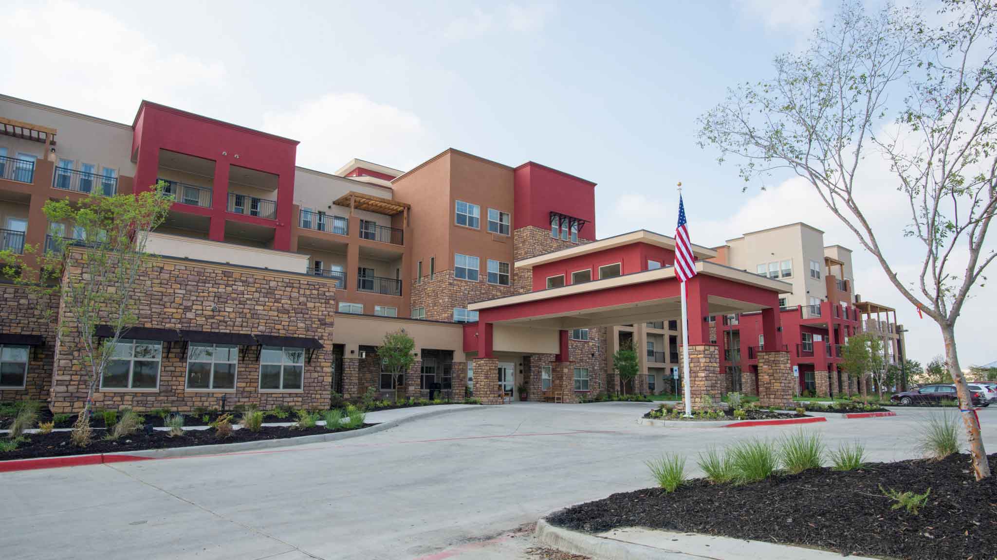 A modern multi-story senior living facility with stone and stucco exterior, an American flag at the entrance, landscaped grounds, and a driveway leading to a covered drop-off area.