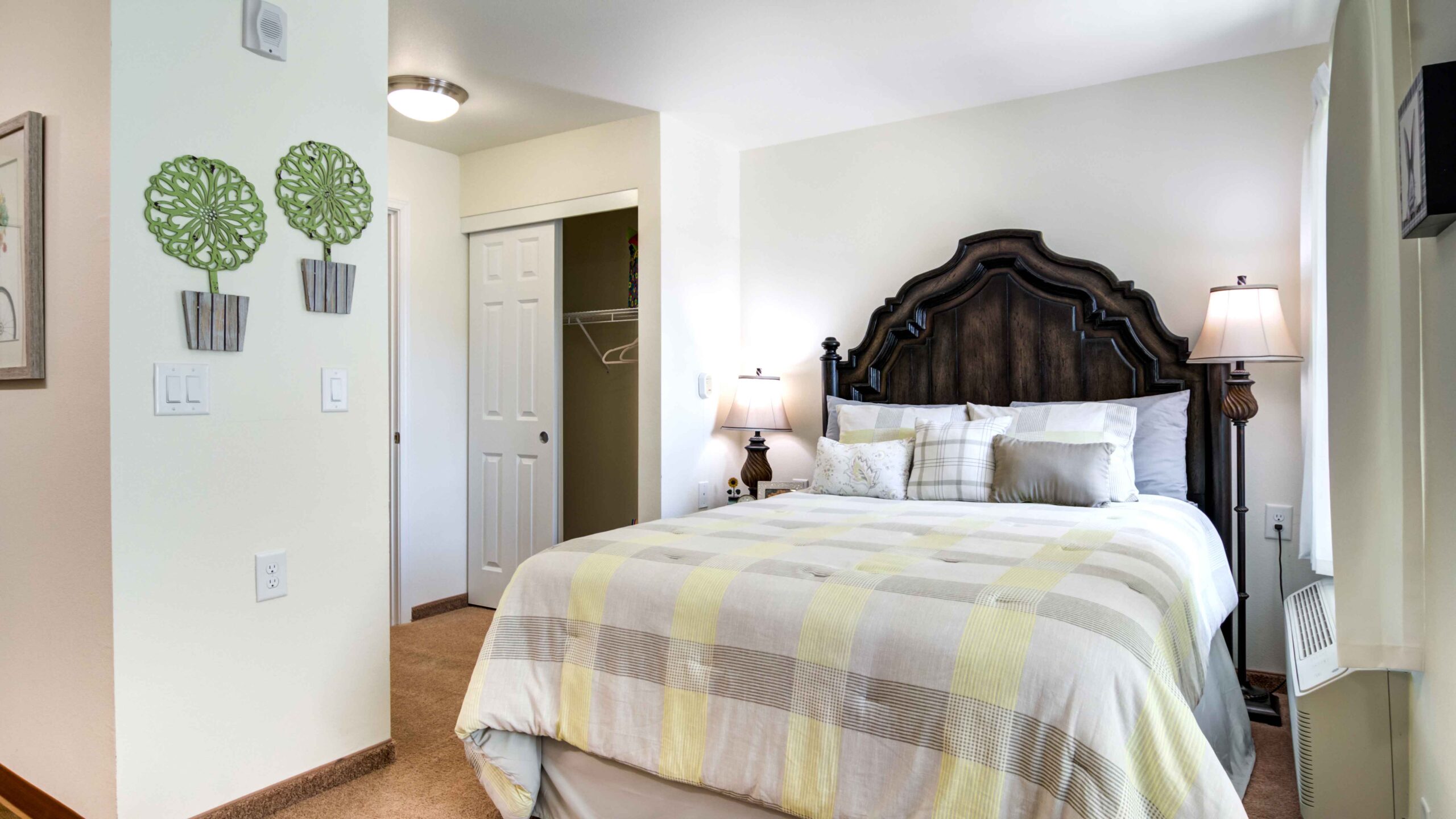 A bright, cozy bedroom with a dark wooden headboard, neatly made bed with pastel striped bedding, two bedside lamps, wall art with green decorative trees, and an open closet in the corner.