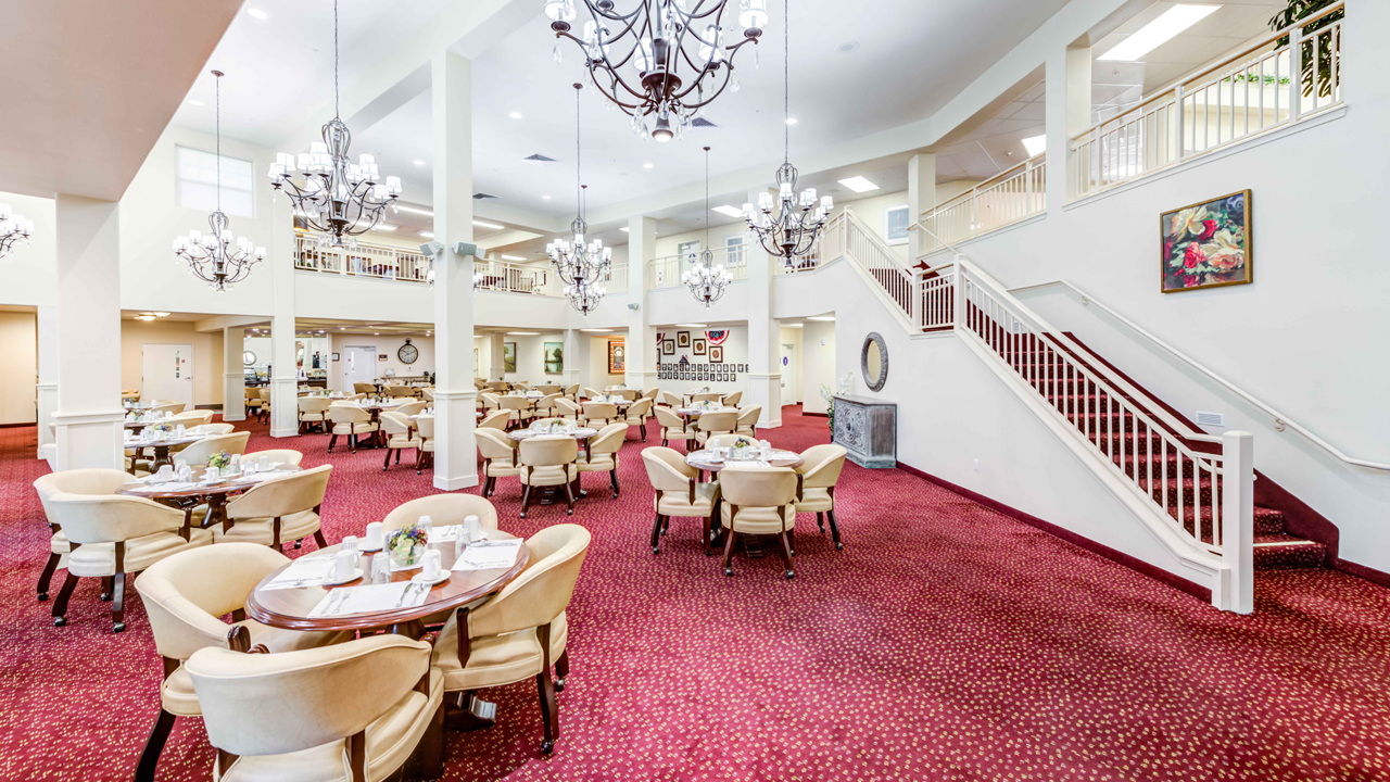 Spacious dining area with red carpet, cream-colored chairs and tables, chandeliers, and a staircase leading to an upper level. The room is brightly lit and elegantly decorated with artwork on the walls.