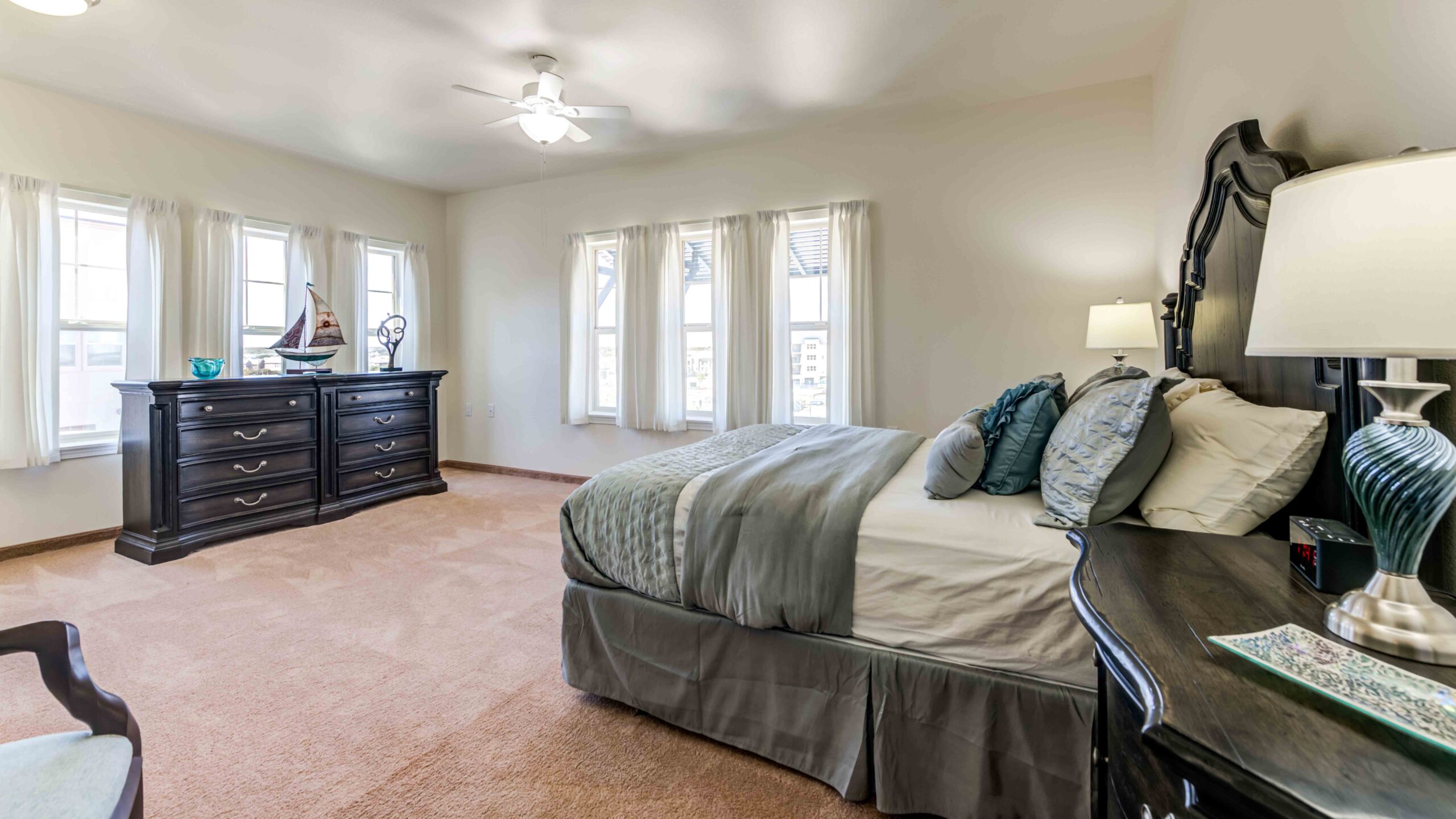 A spacious bedroom with beige carpet, a large bed with gray and white bedding, dark wood furniture including a dresser and nightstand, several windows with white curtains, and a ceiling fan.