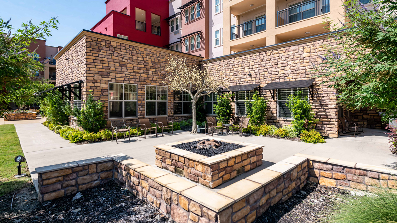 A courtyard with patio chairs around a square stone fire pit, surrounded by stone buildings with balconies, green shrubs, and a small tree on a sunny day.