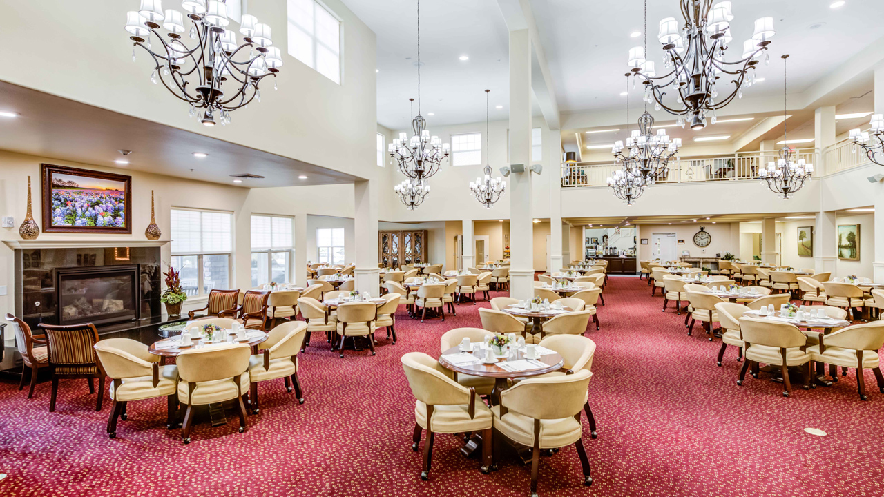 Spacious, elegant dining room with round tables and cream chairs on a red carpet, set for a meal. Large windows, chandeliers, and framed art create a bright, welcoming atmosphere.