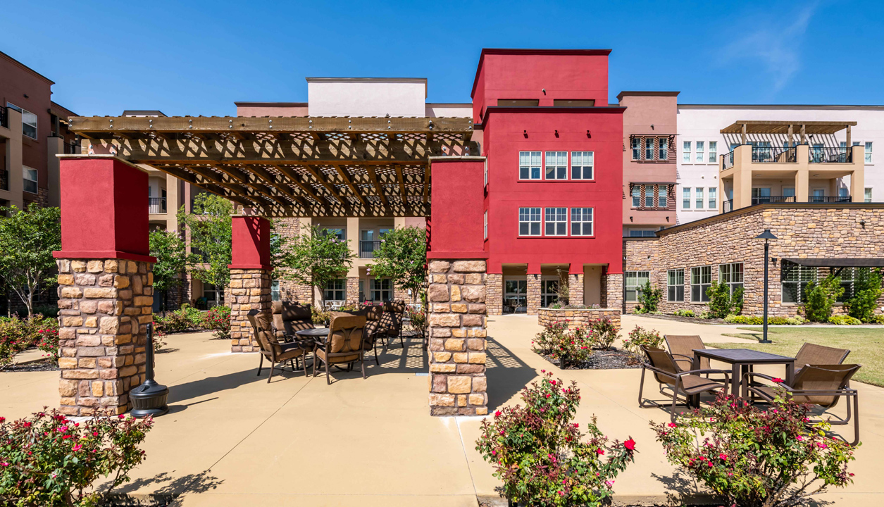 A sunny courtyard of a modern apartment complex features a wooden pergola with seating, surrounded by stone pillars, flowering bushes, and additional patio tables with chairs. The building has red, beige, and stone exterior walls.