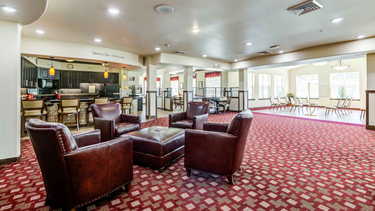 Spacious lounge with four brown leather chairs around a matching ottoman on a red patterned carpet, a counter with barstools, and large windows letting in natural light.