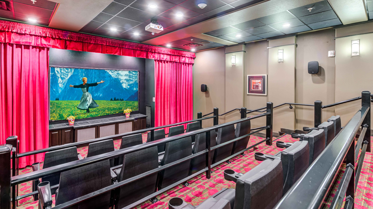 A small home theater with tiered seating, red carpet, and red curtains framing a large screen showing a scene from "The Sound of Music." Walls are beige, and framed art decorates the space. Ceiling lights illuminate the room.