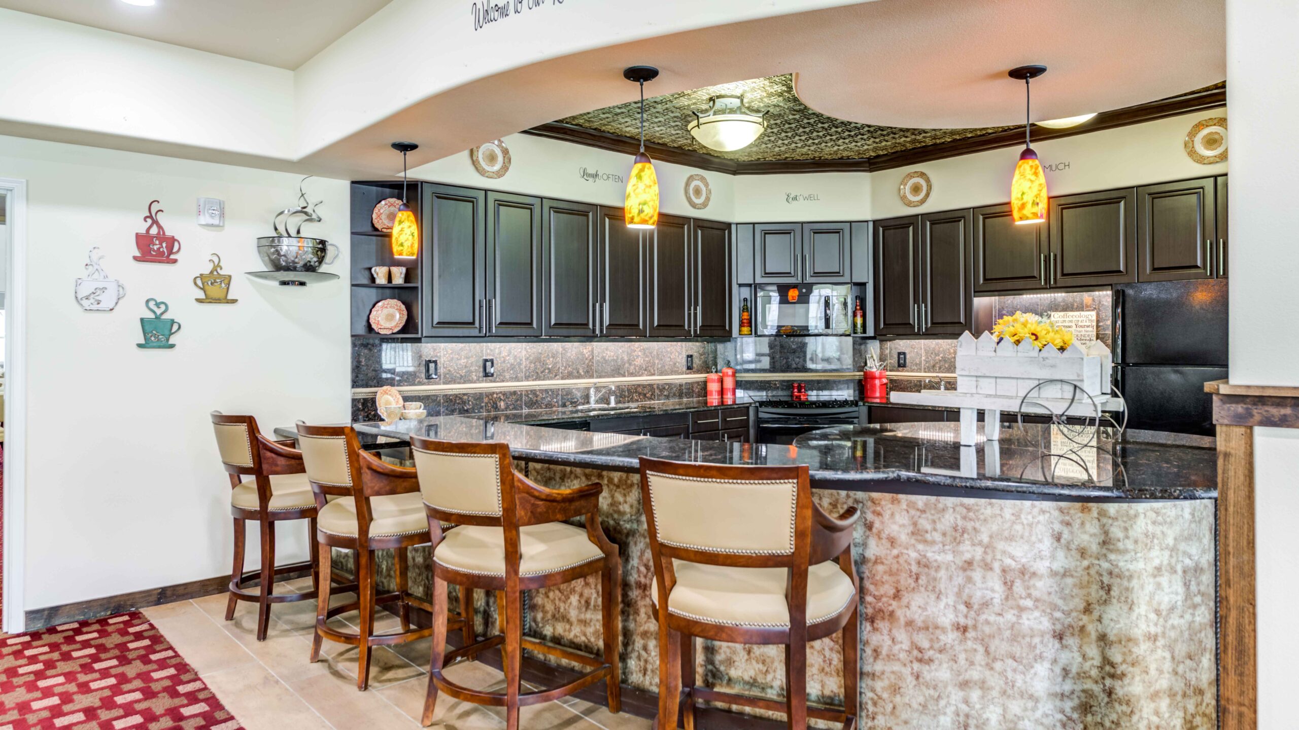A modern kitchen with dark cabinets, a black granite countertop, four upholstered bar stools, hanging pendant lights, and coffee-themed wall decor. Yellow flowers sit on the counter beside decorative jars and baskets.