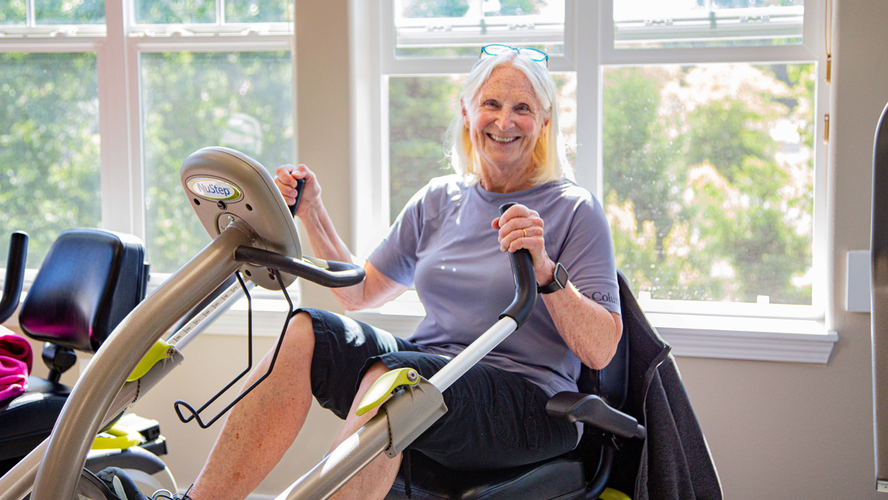 An older woman smiles while exercising on a recumbent cross trainer in a bright room with large windows. She wears a gray shirt, black shorts, and glasses resting on her head.