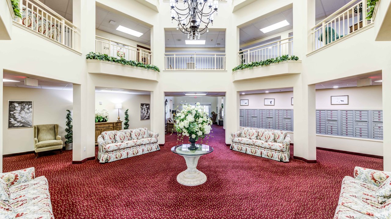 A spacious, bright lobby with red carpet, floral couches, a central round table with a flower arrangement, balconies above, mailbox units on the right, and decorative greenery on the railings.