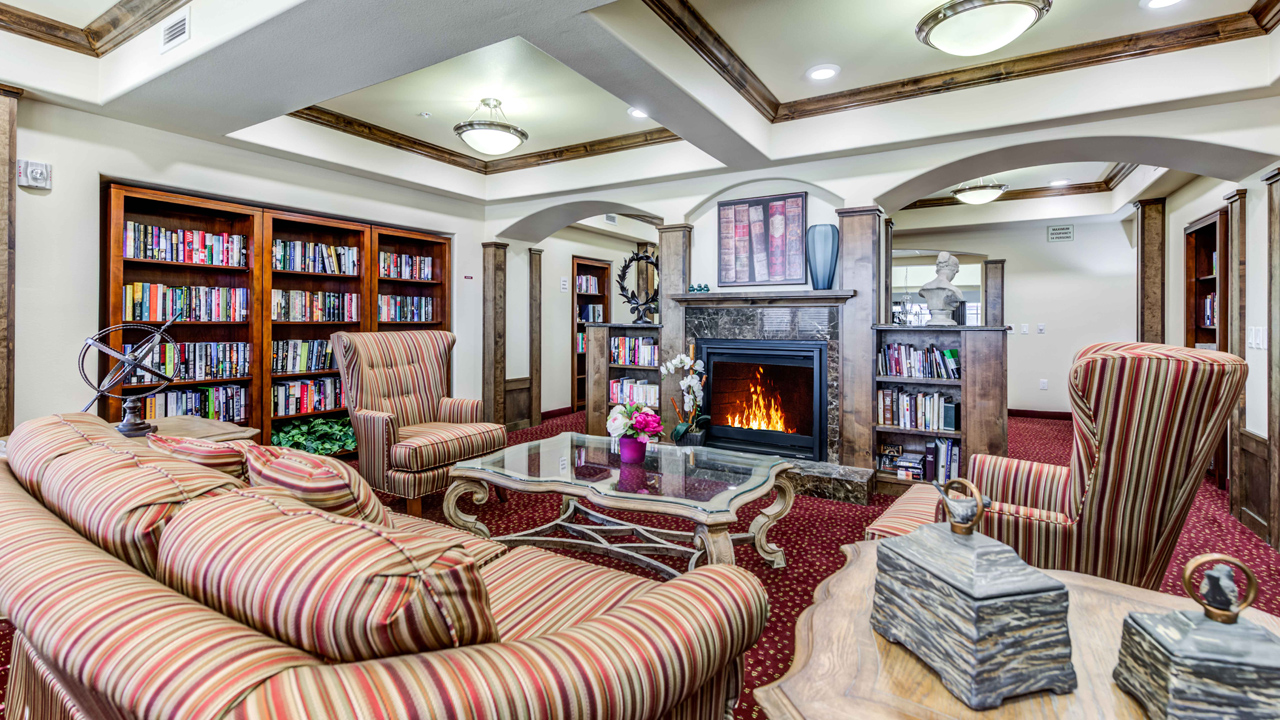 A cozy library with striped armchairs and a sofa around a glass coffee table, a lit fireplace, bookshelves filled with books, red carpet, and elegant decor under soft ceiling lights.