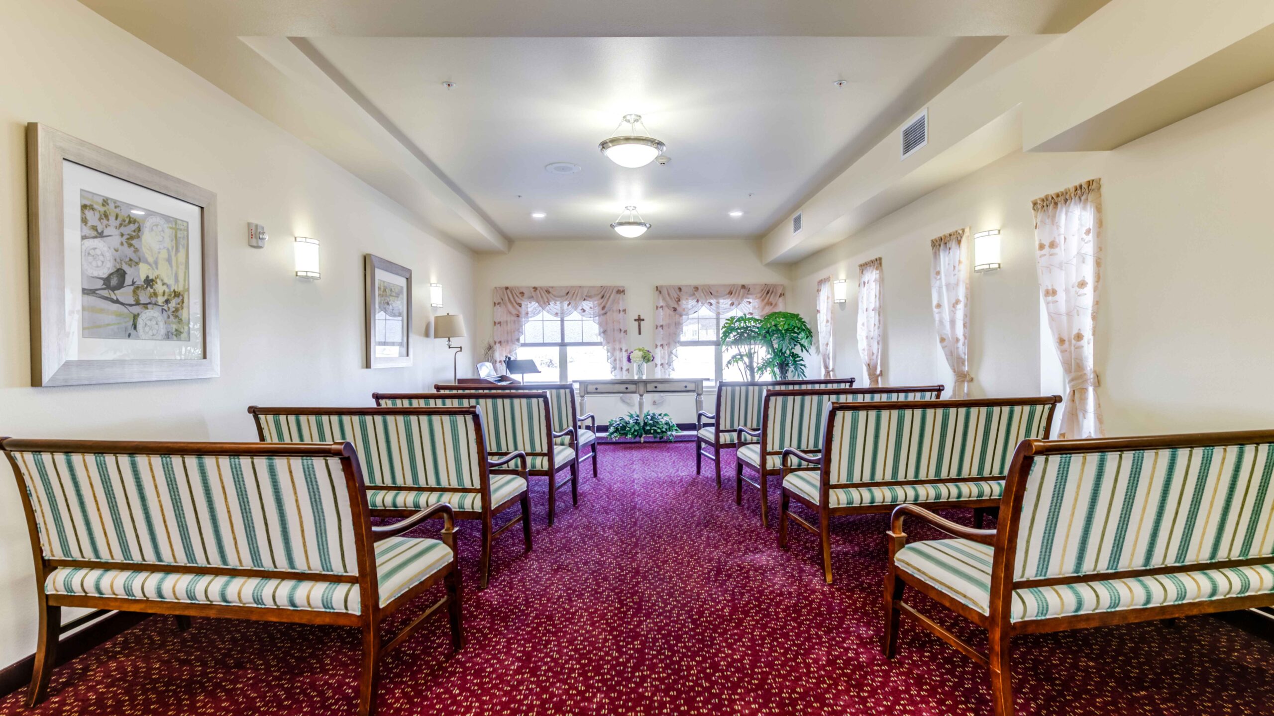 A small chapel with striped wooden chairs arranged in rows facing a window with curtains, cream-colored walls, framed art, plants, and soft ceiling lighting.