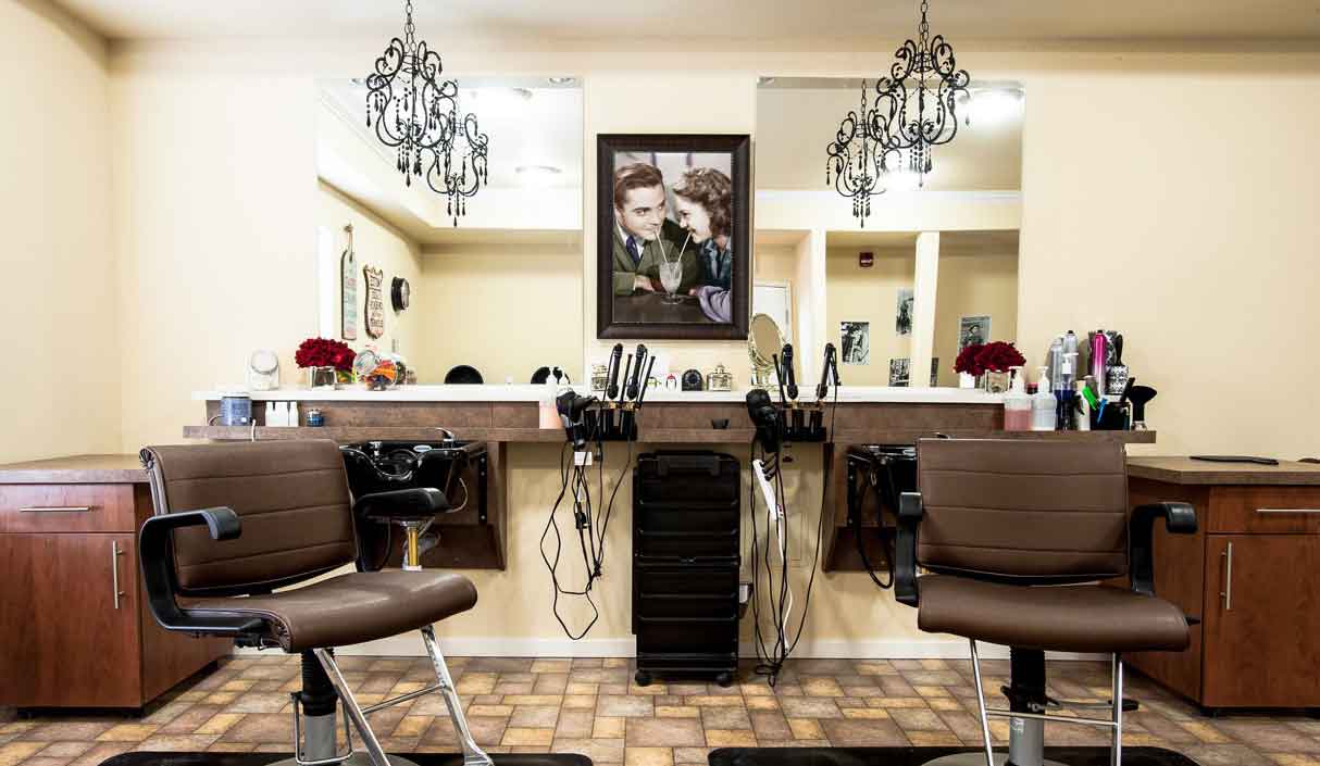 A well-lit hair salon with two brown salon chairs, mirrors, hanging chandeliers, hair tools on the counter, and a framed portrait of two people above the stations.