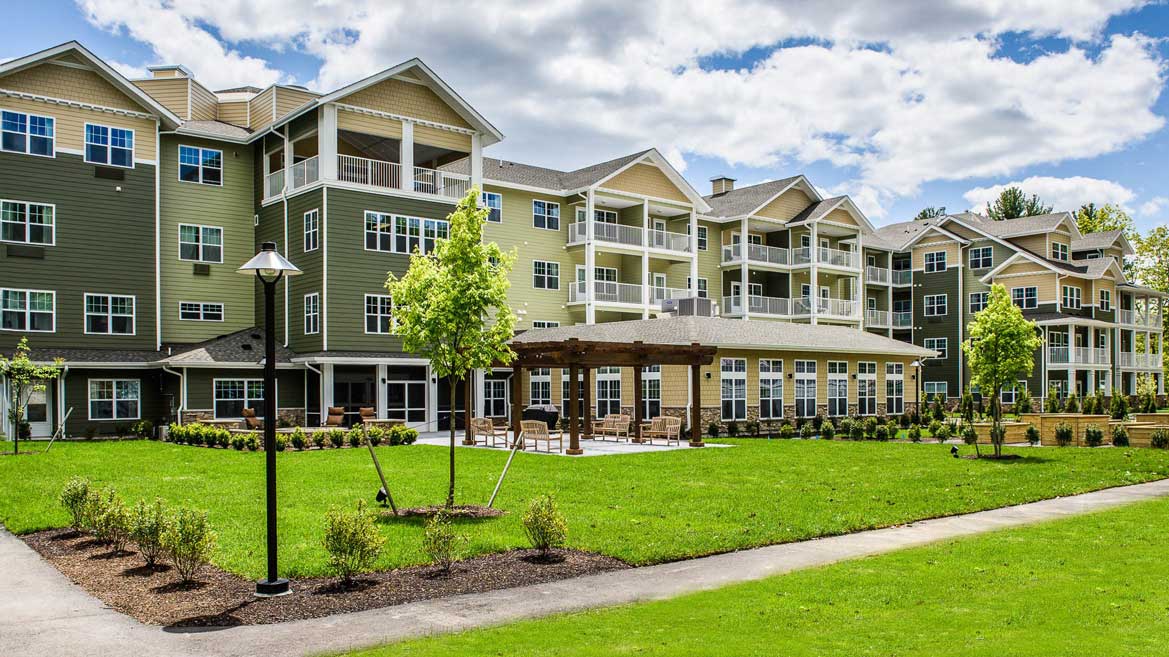 A modern multi-story senior living residential building with green and beige siding, multiple balconies, and large windows. A well-maintained lawn and garden area with a wooden pergola and benches are in the foreground, under a partly cloudy sky.