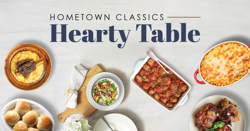 Overhead view of a table with savory dishes including meatballs, mac and cheese, a salad, baked chicken, cornbread, and bread rolls—perfect for Senior Living communities—accompanied by the text "Hometown Classics Hearty Table" above.