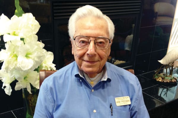 An elderly man with white hair and glasses is sitting in front of a fireplace, wearing a blue shirt with a name tag. White flowers are visible to his left.