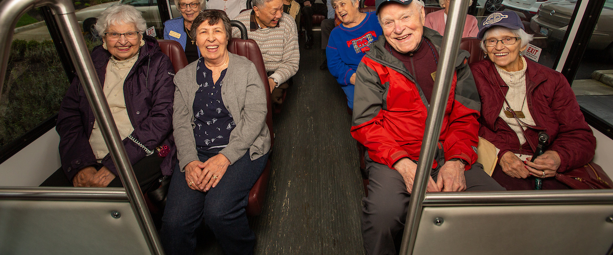 A group of elderly people is seated on a bus, smiling and enjoying a ride. They are casually dressed, and some are chatting with each other. The atmosphere appears cheerful and friendly. The bus seats are maroon, and the interior looks well-lit.