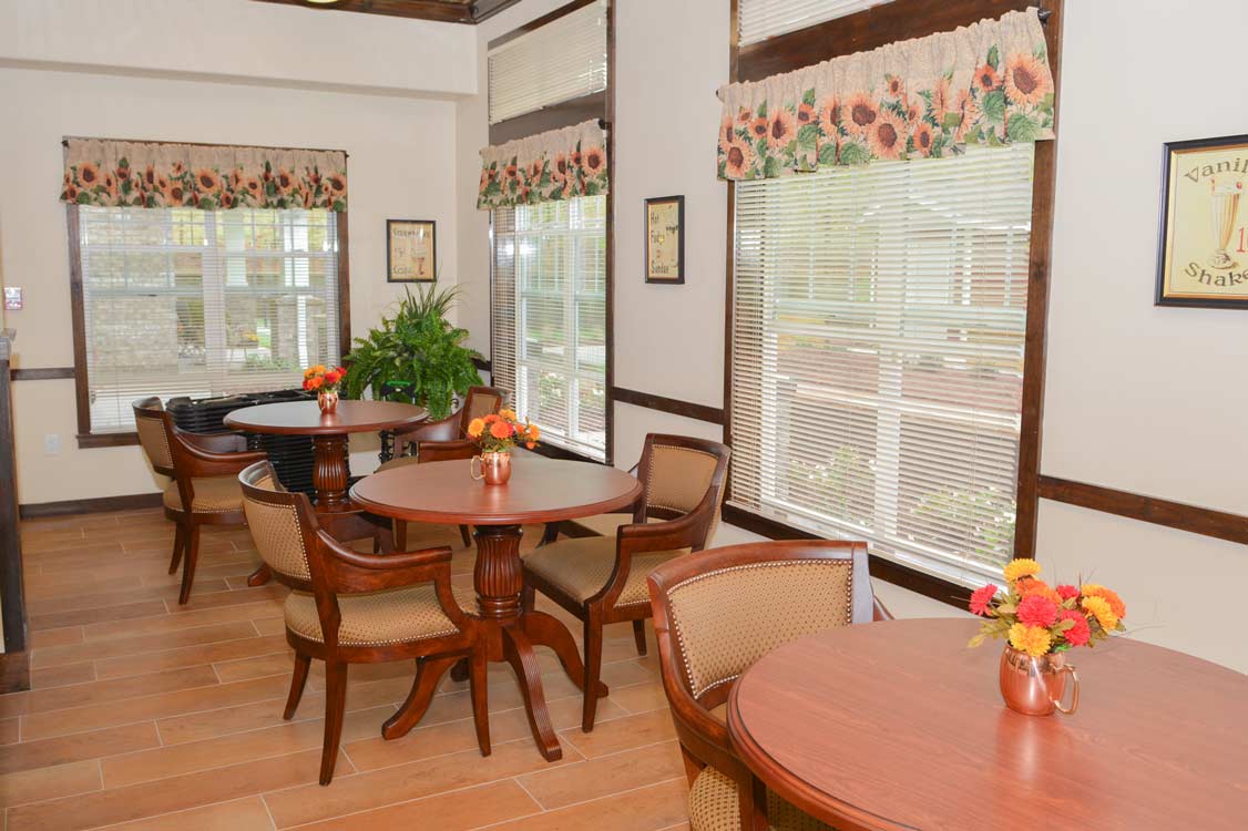 A cozy cafe with several round wooden tables and cushioned chairs. The tables have flower arrangements in copper-colored vases. Large windows decorated with sunflower-themed valances let in natural light, and framed artwork adorns the walls.