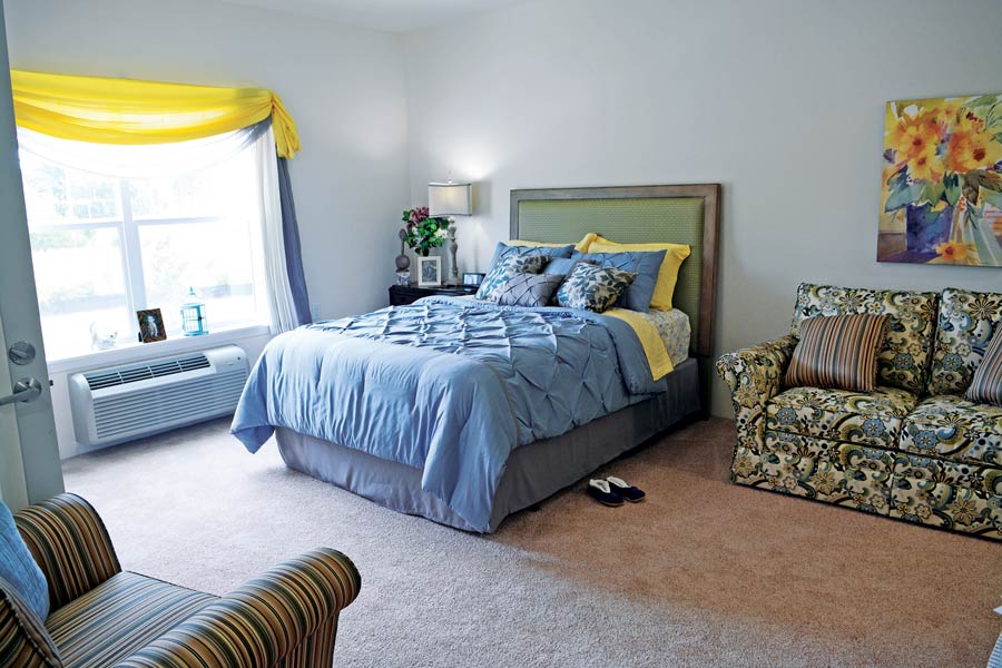 A cozy bedroom featuring a bed with blue and yellow bedding, flanked by a nightstand with a lamp. A window with yellow curtains is on the left side of the room. There's a floral sofa and a striped armchair, as well as a colorful flower painting on the wall.