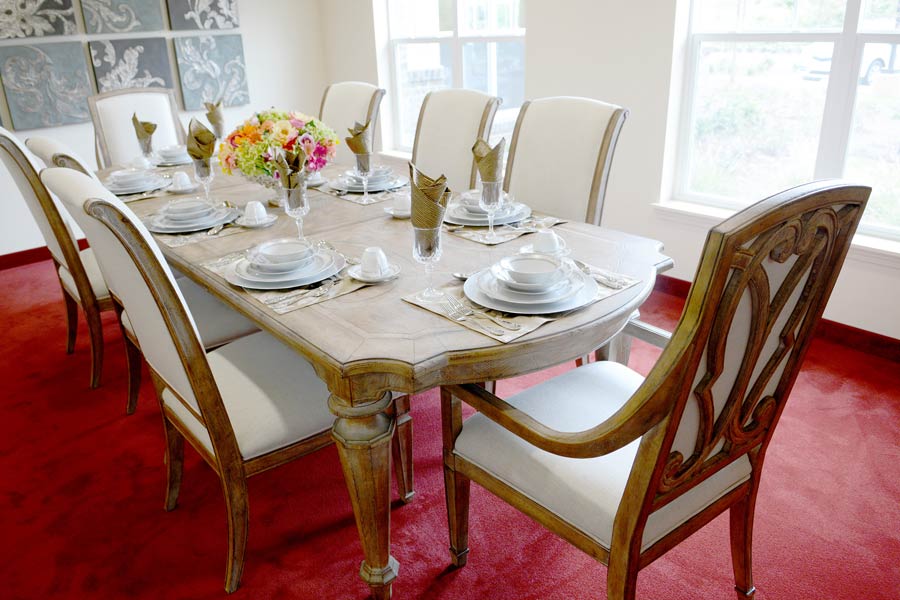 A dining room with a wooden table set for eight people, featuring white cushioned chairs. The table is adorned with a floral centerpiece and each setting includes white plates, bowls, and cups on beige placemats with folded napkins. The room has red carpet flooring.