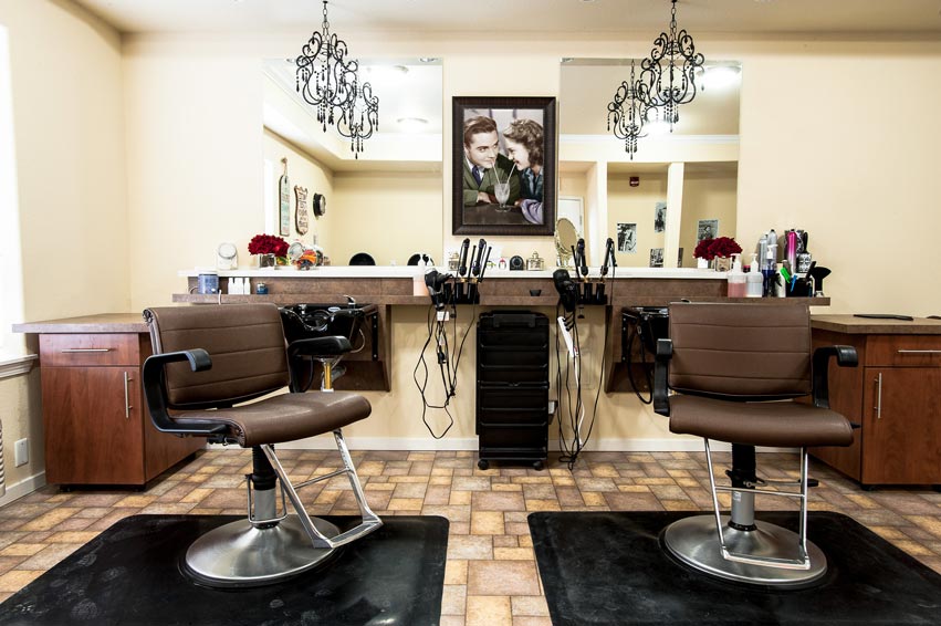 A well-lit hair salon features two brown leather styling chairs on black mats facing a mirror-lined wall. Above the chairs are ornate chandeliers. On the counter behind the chairs are various hair styling tools and products, with a framed picture on the wall.