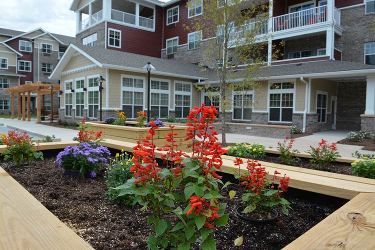 A courtyard garden features vibrant red and purple flowers in raised wooden planters. The background shows a multi-story residential building with large windows, balconies, and red and beige siding. The courtyard has pathways and lampposts.