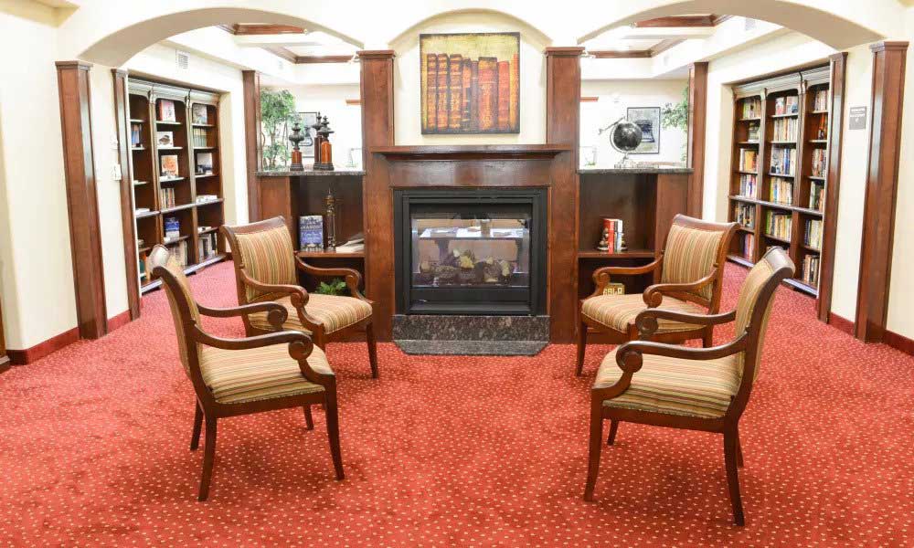 A cozy library interior with a red carpeted floor. Four wooden chairs with striped cushions are arranged in a circle around a central fireplace with a mantel. Bookshelves filled with books line the walls. A painting of books is displayed above the fireplace.