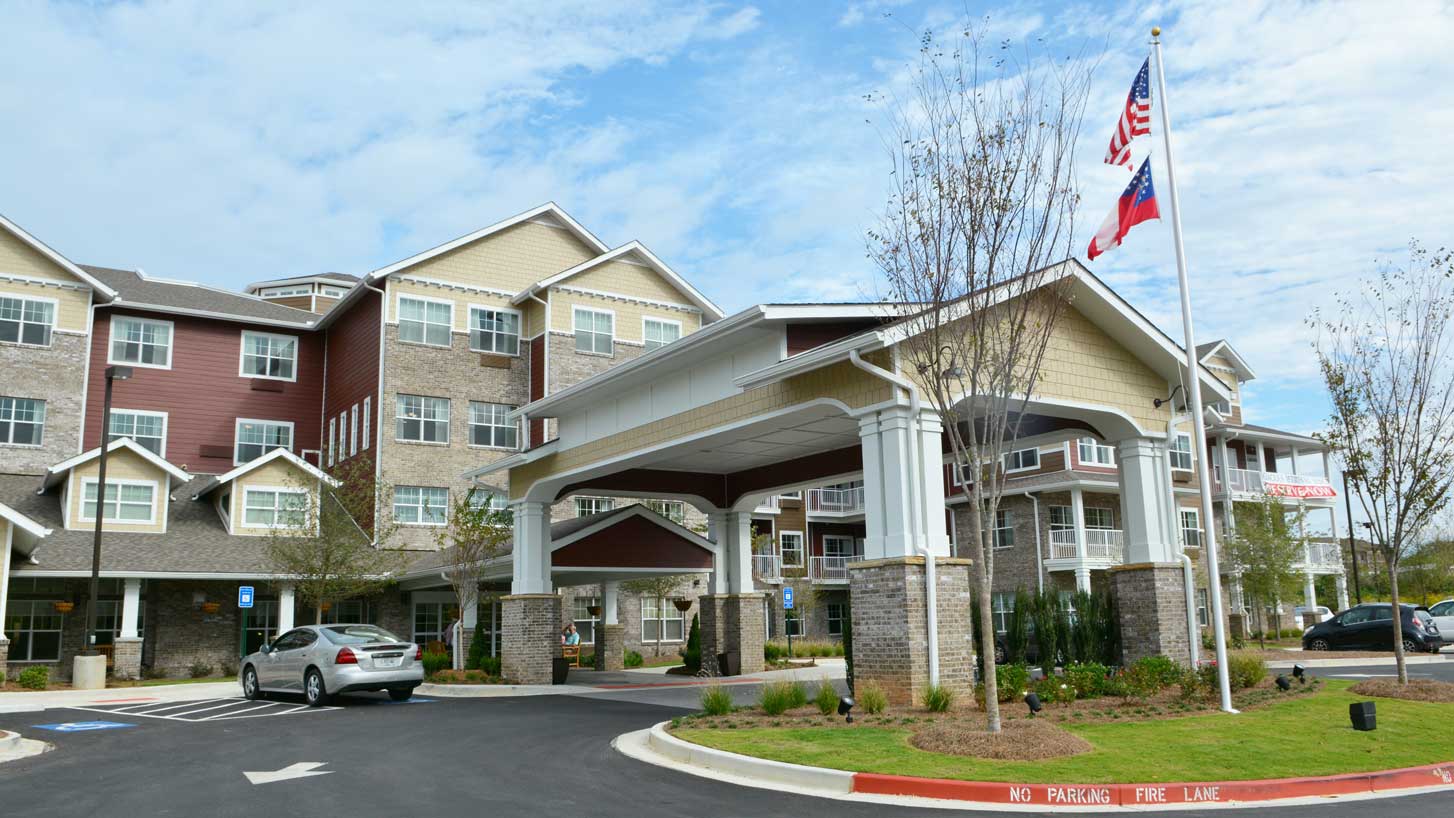 A multi-story assisted living facility with a covered entrance and a circular driveway. Two flags are flying on poles near the entrance: the American flag and the state flag. A parked car is visible in front of the building, and landscaped greenery surrounds the area.