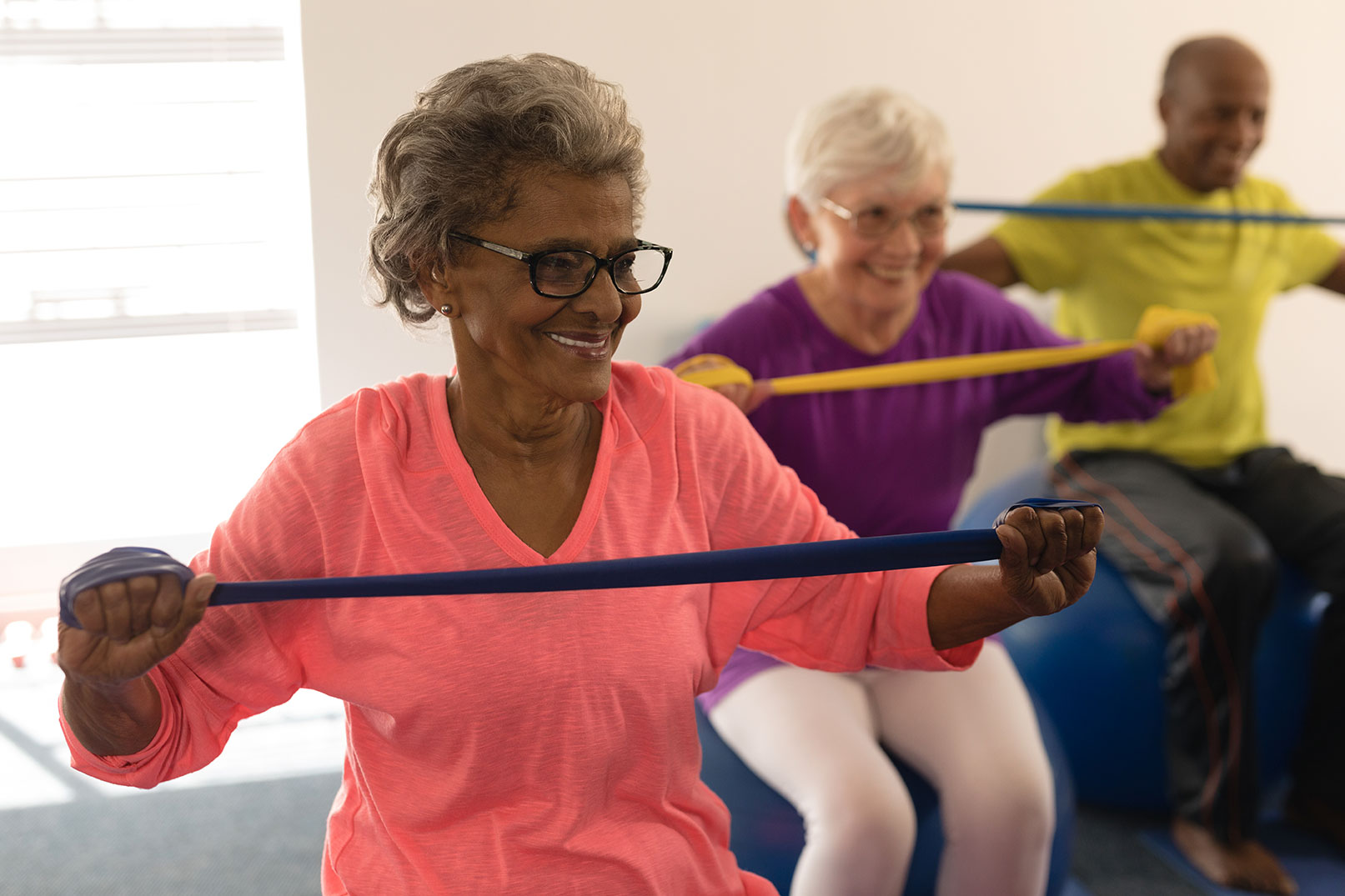 Three older adults are participating in a seated exercise class, using resistance bands. A woman in a pink top is in the forefront, smiling. Behind her, a woman in purple and a man in yellow are exercising as well. They all appear engaged and cheerful.