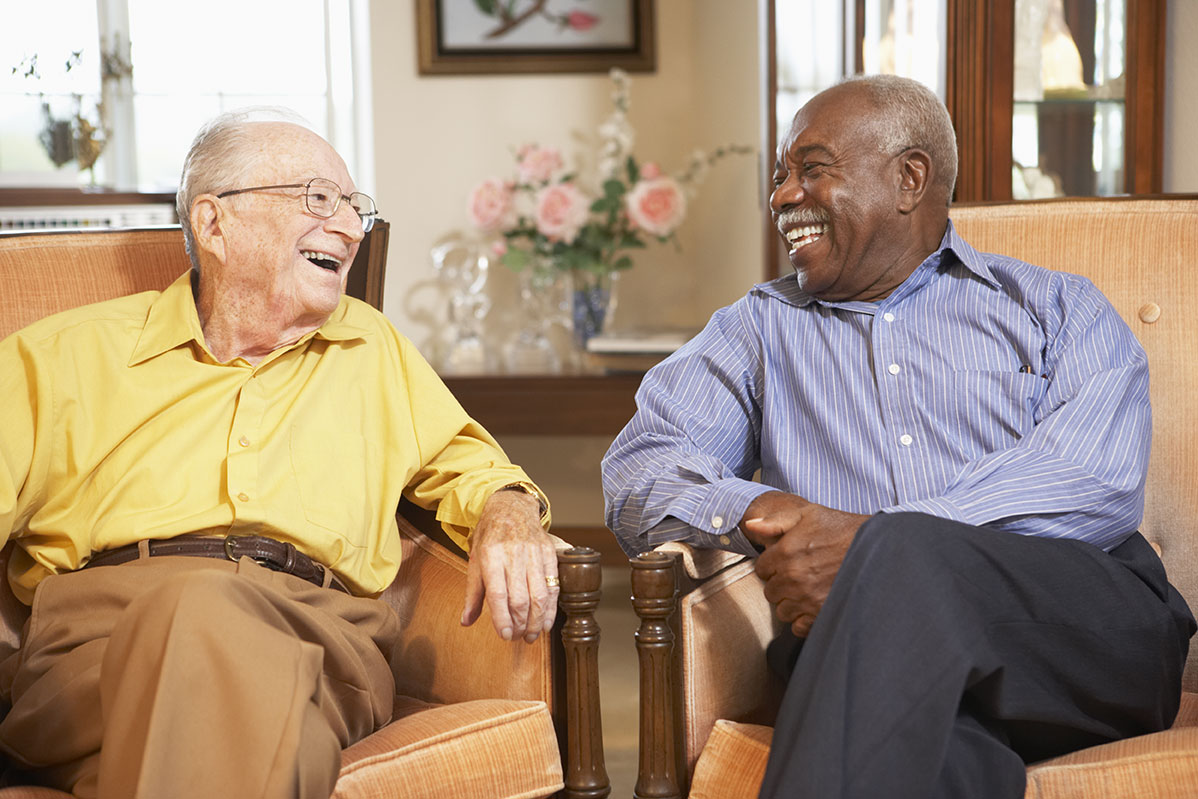 Two elderly men sit in comfortable armchairs, smiling and laughing together. One wears a yellow shirt and brown pants, while the other wears a blue striped shirt and dark pants. They appear to be in a warmly lit living room with flowers and decorations in the background.