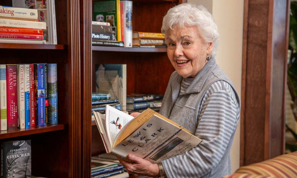 An elderly woman with white hair smiles while holding and looking at a book titled 