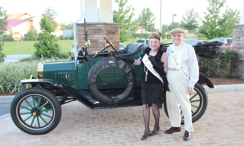 A smiling couple stands next to a vintage green car. The woman, in a flapper dress and black feathered headband, wears a 