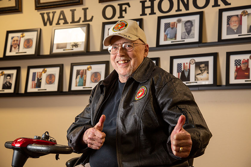 An elderly man wearing a leather jacket and a cap sits in a mobility scooter, smiling and giving two thumbs up. Behind him, a 