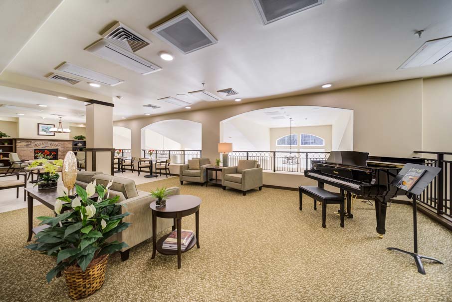 A spacious lounge area with beige carpet and light-colored walls features several seating arrangements, including sofas and armchairs around a table with a lamp. A grand piano with sheet music on a stand sits nearby, and potted plants decorate the space. Large arched windows allow natural light to fill the room.