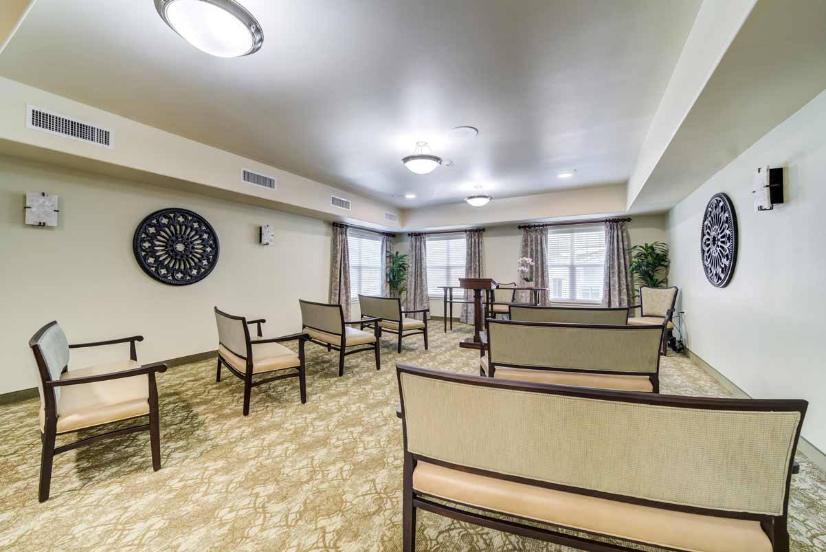 A small, well-lit room with beige walls and carpet, containing several cushioned chairs arranged in rows facing a podium at the front. Large decorative wall art and plants are placed along the walls, and windows with curtains let in natural light.