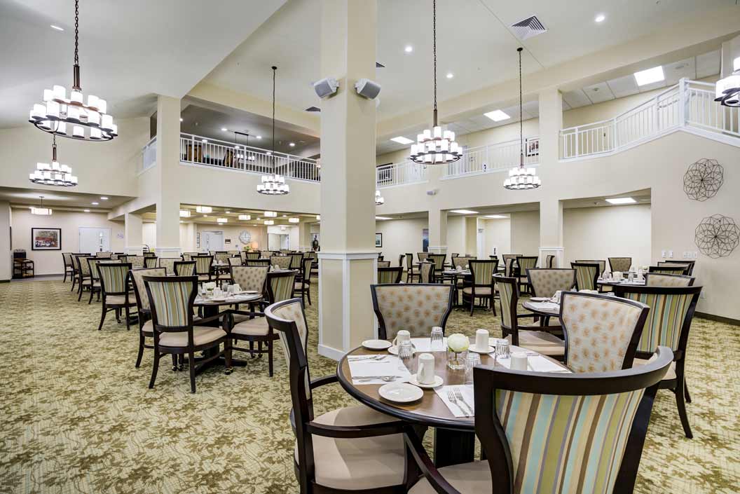 A spacious, elegantly decorated dining room with round tables set with white dishes and coffee cups. The high ceiling features chandeliers, and there is an upper level with railings. The room has a patterned carpet and walls adorned with simple artwork.