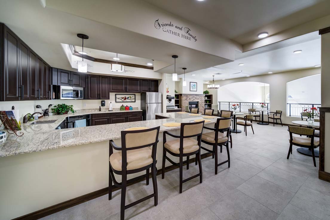 A modern kitchen and dining area with granite countertops, wooden cabinets, and bar stools. The space also features a stainless steel fridge, a microwave, and multiple dining tables with chairs. A decorative sign reads 