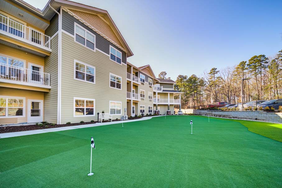 A beige multi-story building with balconies overlooks a well-maintained green lawn equipped with putting greens for golfing. Trees are visible in the background, and the scene is under a clear blue sky.