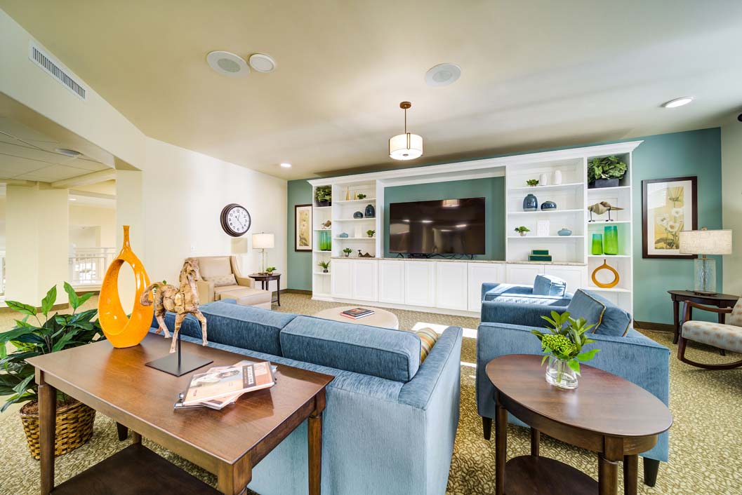 A modern living room with a large flat-screen TV mounted on a white built-in entertainment center. The room features a blue sectional sofa, wooden coffee and side tables, decorative items, and a clock on the wall. The floor is carpeted, and the room is well-lit.