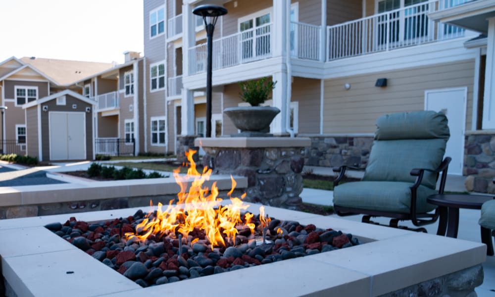 A modern apartment complex with balconies and a stone facade, featuring a cozy outdoor fire pit with flames and surrounding chairs. The area is furnished with patio furniture, and a small shed is visible in the background.