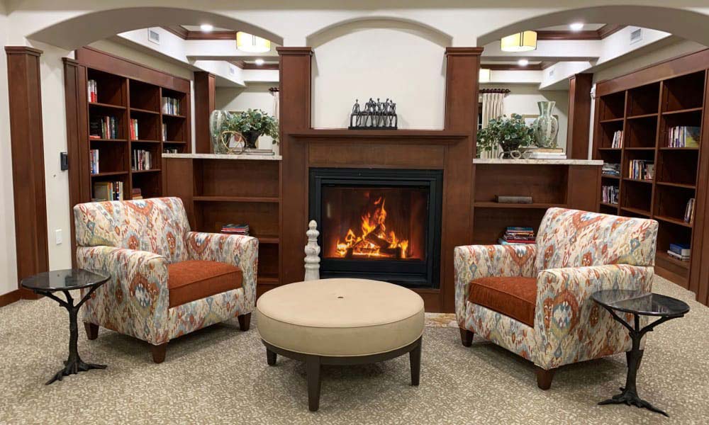 A cozy room features a lit fireplace centered between two wooden bookshelves filled with books and decor. In front of the fireplace, two patterned armchairs with brown cushions flank a round ottoman. Small side tables are placed next to each chair.