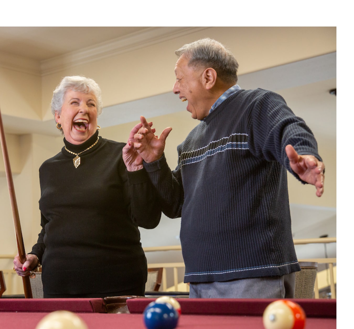 An elderly woman and man share a laugh while standing near a pool table. The woman holds a pool cue and wears a black sweater with a necklace, while the man, in a black-and-gray sweater, gestures animatedly. Pool balls are visible on the table in front of them.