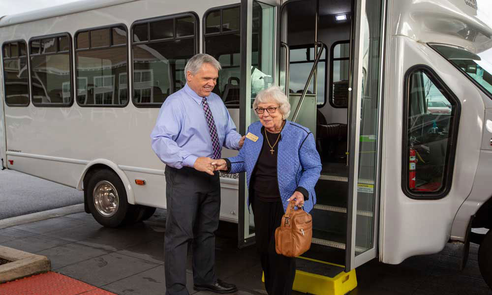 A man in a blue shirt assists an elderly woman with white hair, glasses, and a light blue coat as she steps off a shuttle bus. He is holding her hand and offering support. The woman carries a brown handbag. The shuttle bus has an open door and a small step.
