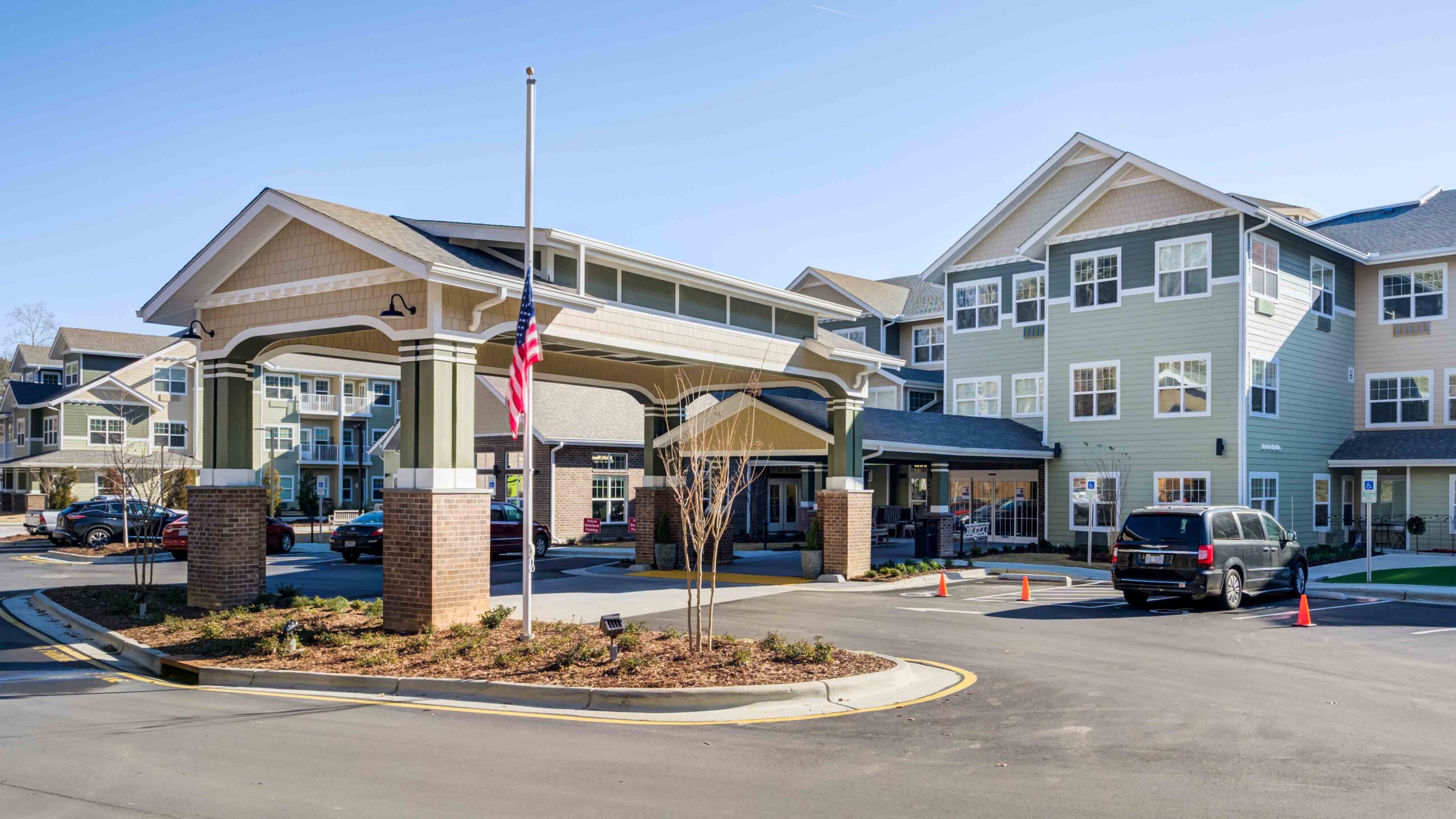 A modern assisted living facility with light green siding, large windows, a covered entrance, and an American flag at half-staff; cars are parked nearby on a sunny day.