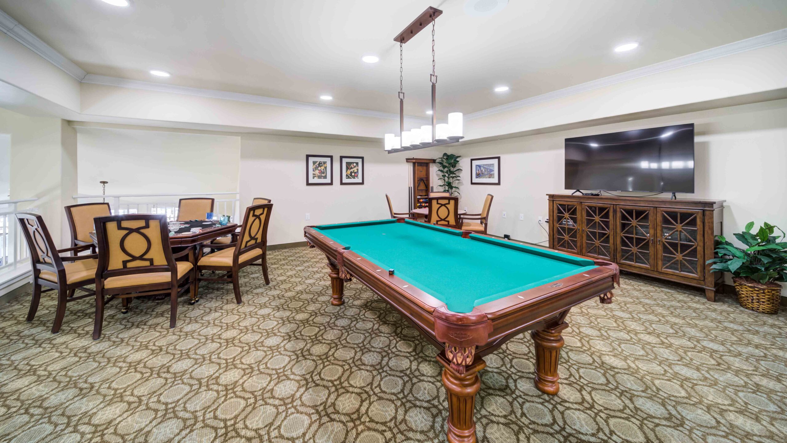 A spacious game room with a pool table in the center, a round table with chairs to the left, a large flat-screen TV on a wooden cabinet to the right, and framed artwork on the back wall.