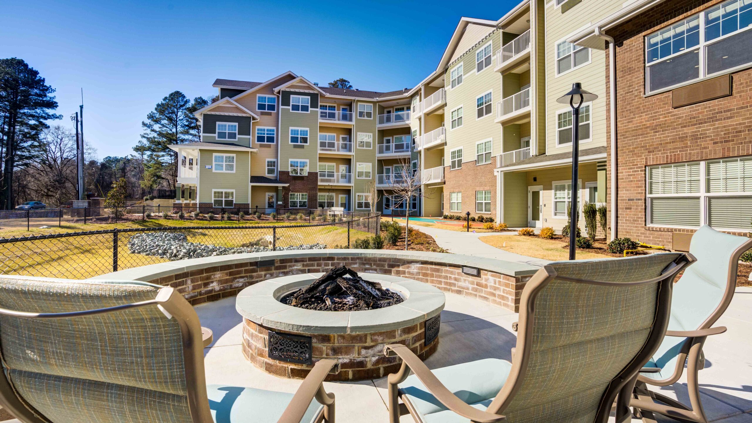 Outdoor seating area with a round fire pit and chairs, set in front of a modern multi-story apartment building with balconies, landscaped grounds, and trees in the background under a clear blue sky.