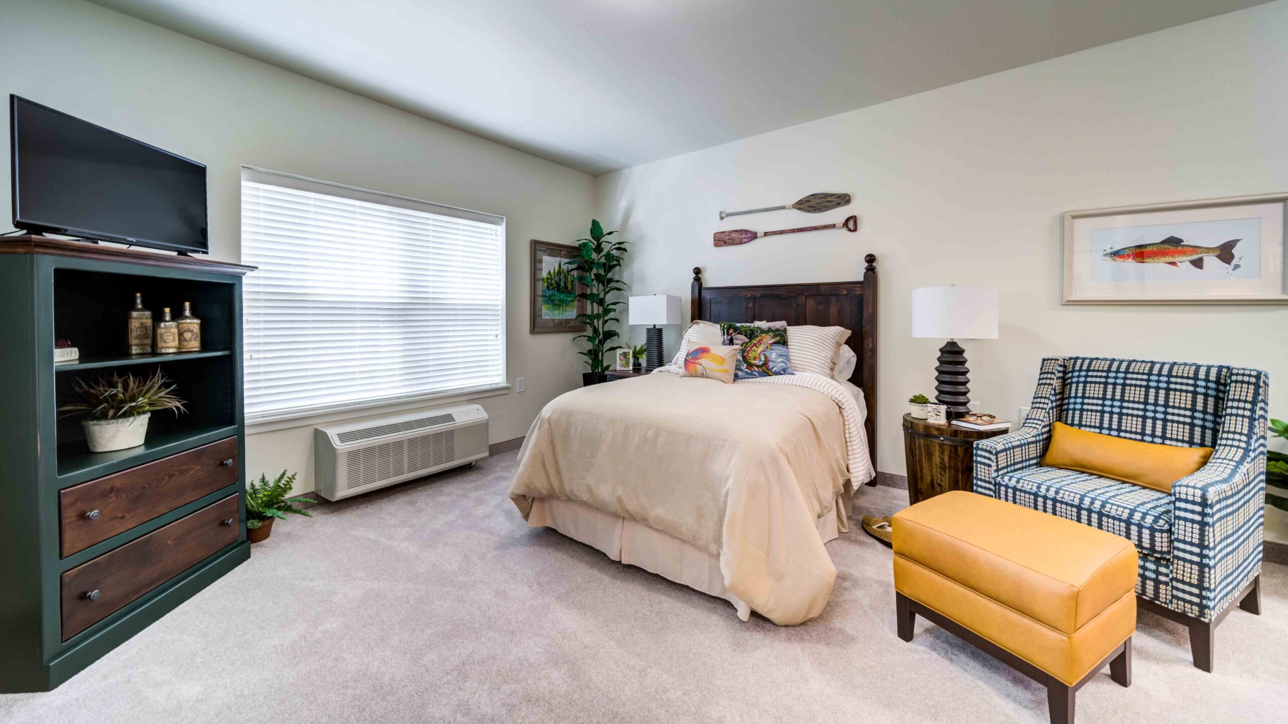 A cozy bedroom with a neatly made bed, plaid armchair with a yellow ottoman, a green dresser with a TV, wall art, a window with blinds, and a potted plant in the corner. The room has carpeted flooring.